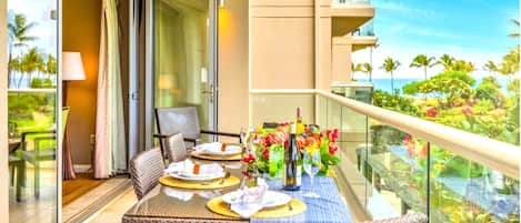 Un balcon avec une table dressée pour le dîner, des chaises en osier et une vue sur les palmiers et l’océan.