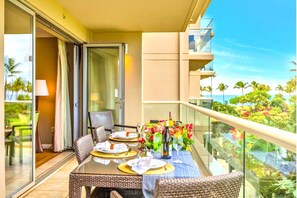 A balcony with a table set for dining, wicker chairs, and a view of palm trees and the ocean.