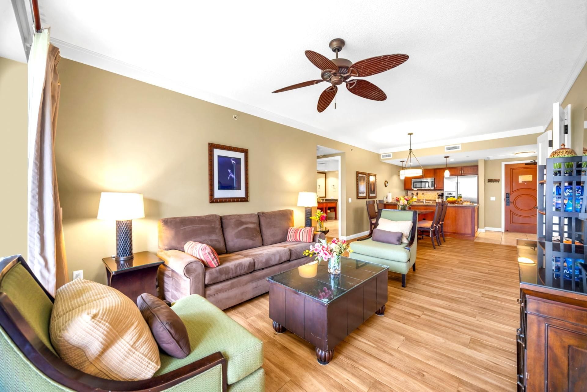 Ein geräumiges Wohnzimmer mit einem braunen Sofa, einem grünen Sessel und einem Couchtisch. Ein Deckenventilator ist sichtbar, und der Boden ist mit Holz ausgelegt.