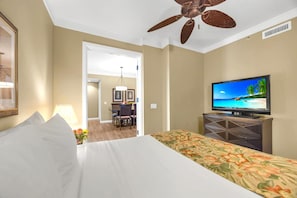 A hotel room with a bed, a television, a ceiling fan, and a dining area in the background.
