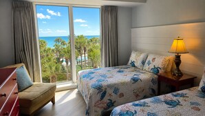 A hotel room with two beds, a nightstand with a lamp, a dresser, and a large window offering a view of the ocean and palm trees.