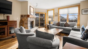 A living room with a stone fireplace, grey sofas, a flat-screen TV, and large windows with blinds. - High-end Finishes, 2 King Beds, Private Laundry, Overlooks Dercum Square (Keystone)