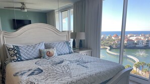 Een slaapkamer met een groot bed, een televisie, een raam met uitzicht op een wateroppervlak en gebouwen, en een plafondventilator.