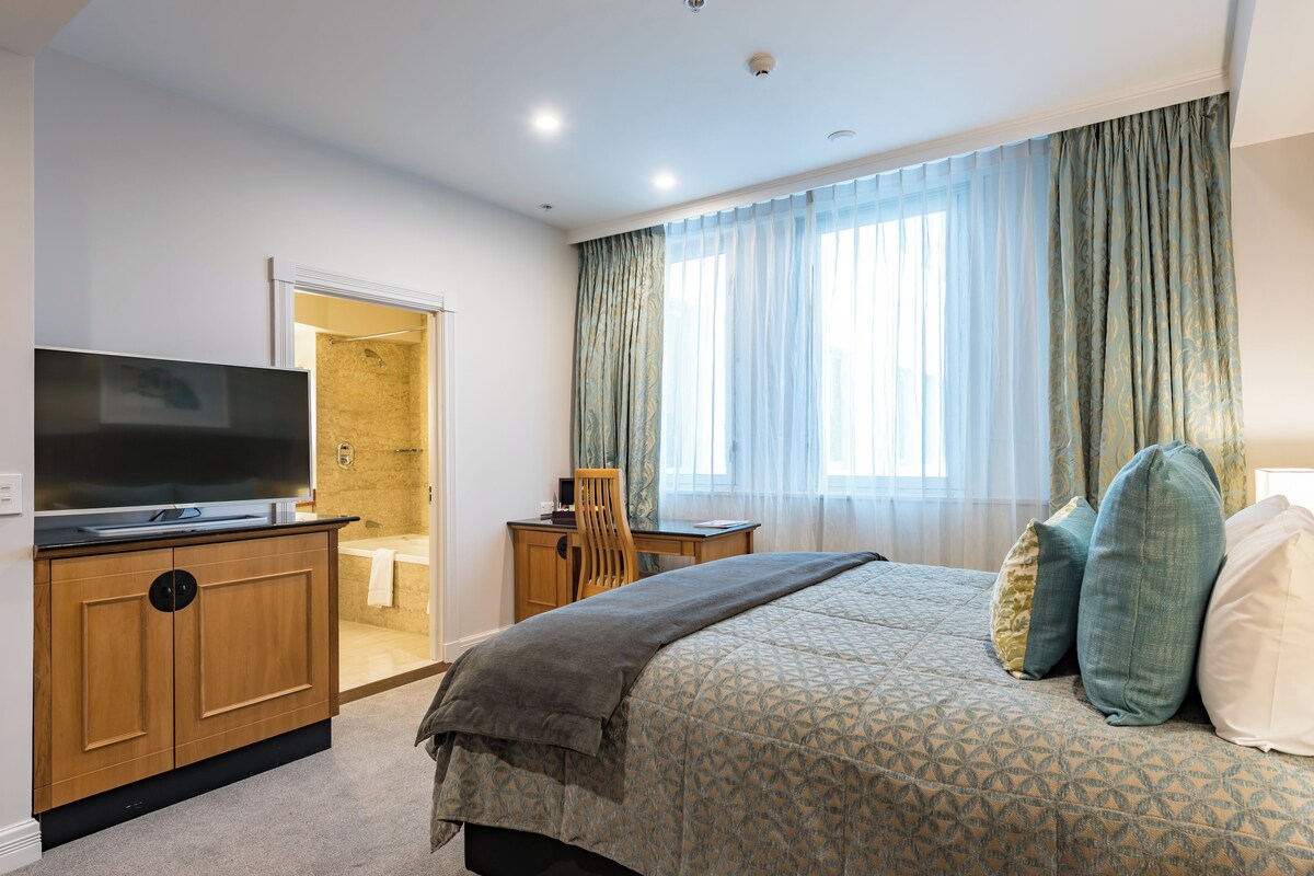 Een slaapkamer met een bed, een televisie op een houten dressoir, een bureau en een badkamer die door een open deur zichtbaar is.
