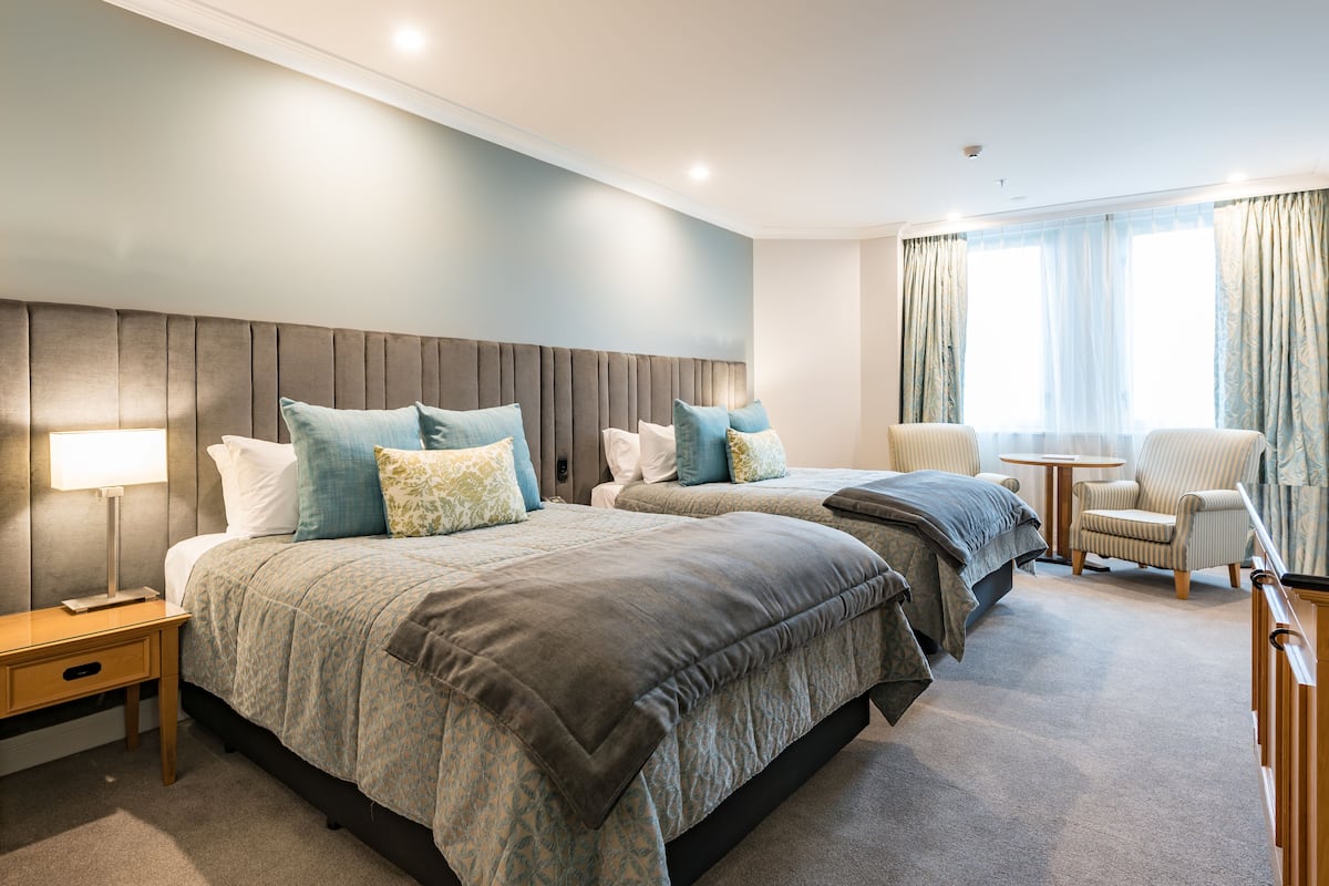 Een hotelkamer met twee bedden, een nachtkastje met een lamp, een stoel en een klein rond tafeltje.