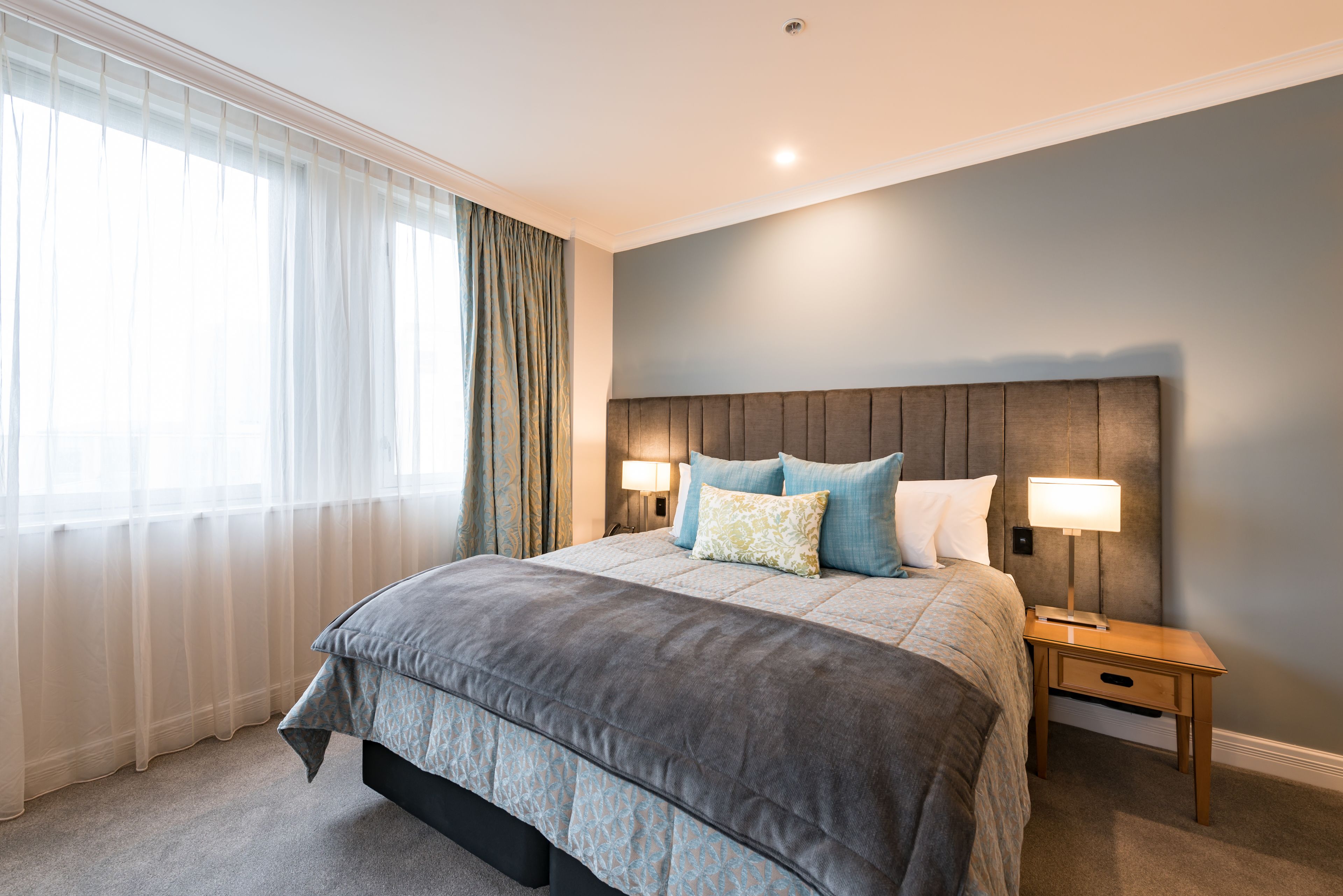 A bedroom with a large bed, two bedside tables with lamps, a window with sheer curtains, and a plain wall.
