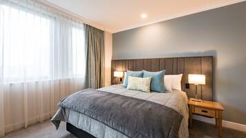 A bedroom with a large bed, two bedside tables with lamps, a window with sheer curtains, and a plain wall.