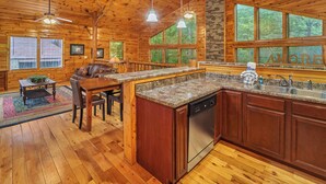 A wooden cabin interior with a kitchen, dining area, and large windows.