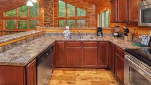 A kitchen with wooden cabinets, granite countertops, a dishwasher, a stove, and a large window overlooking a forest.