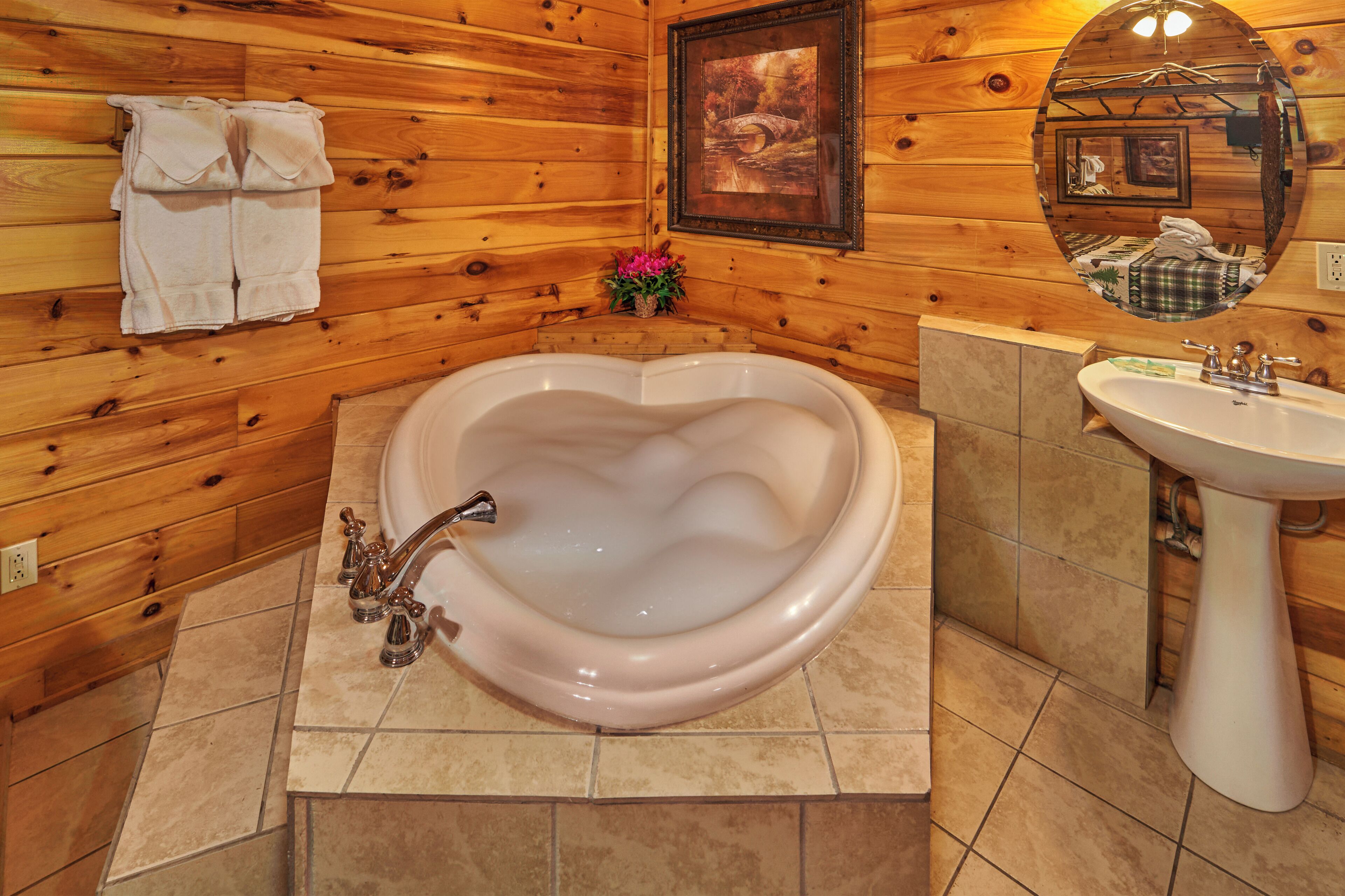 A heart-shaped bathtub in a wooden cabin bathroom with a sink and a framed picture above the mirror.