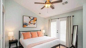 Une chambre à coucher comprenant un lit, des tables de chevet, un ventilateur de plafond, un tableau, une fenêtre avec des rideaux et un miroir.