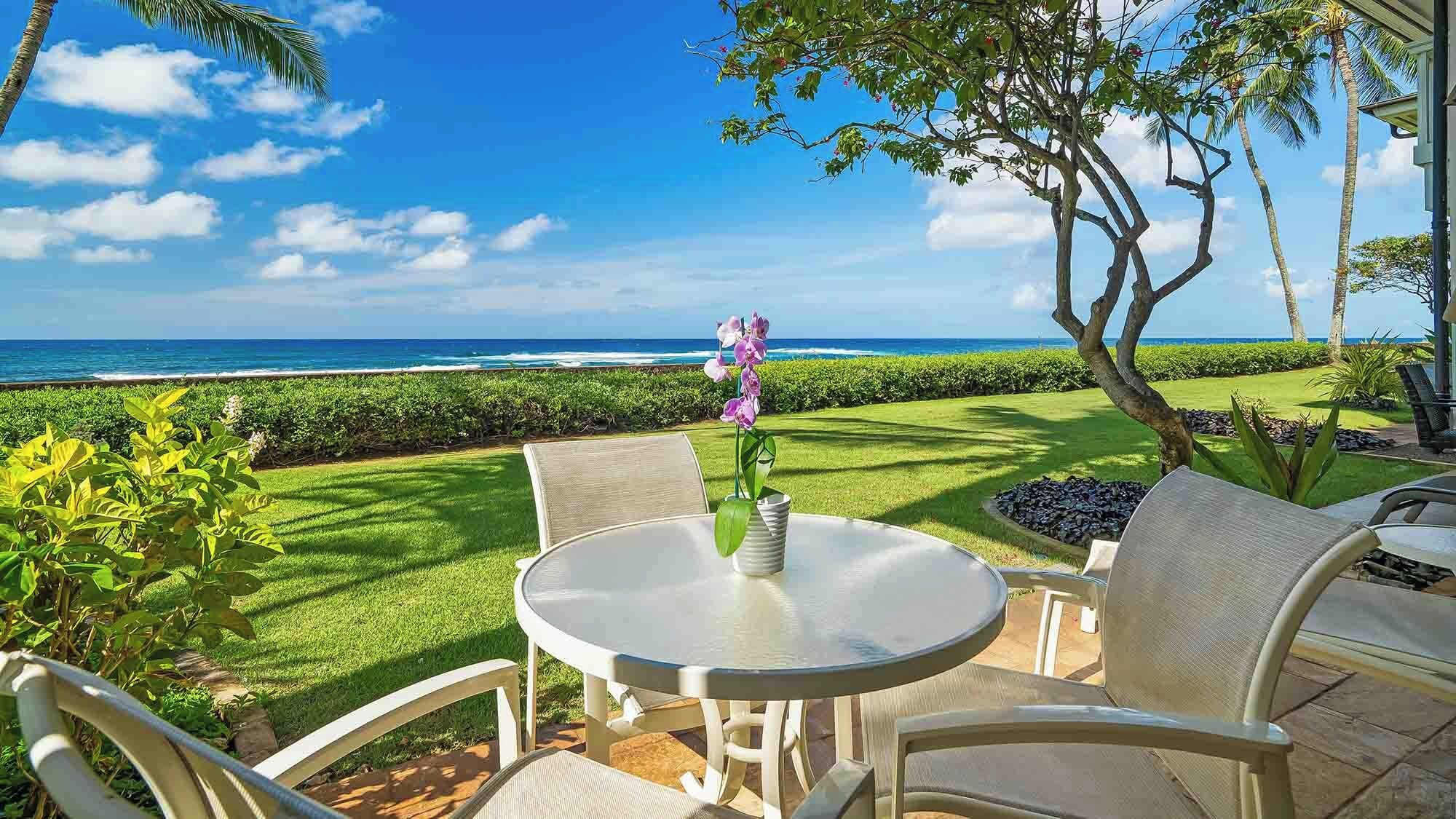 A patio with a table and chairs, a vase with flowers, and a view of the ocean.