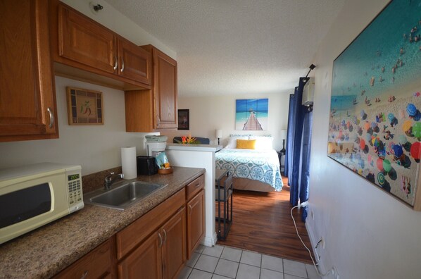 A kitchen with wooden cabinets, a microwave, and a sink. There is a bed with a blue and white patterned bedspread and a painting of a beach scene on the wall.