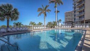 Una piscina azul cristalina rodeada de sillas de descanso blancas y palmeras, con un edificio de varios pisos al fondo.