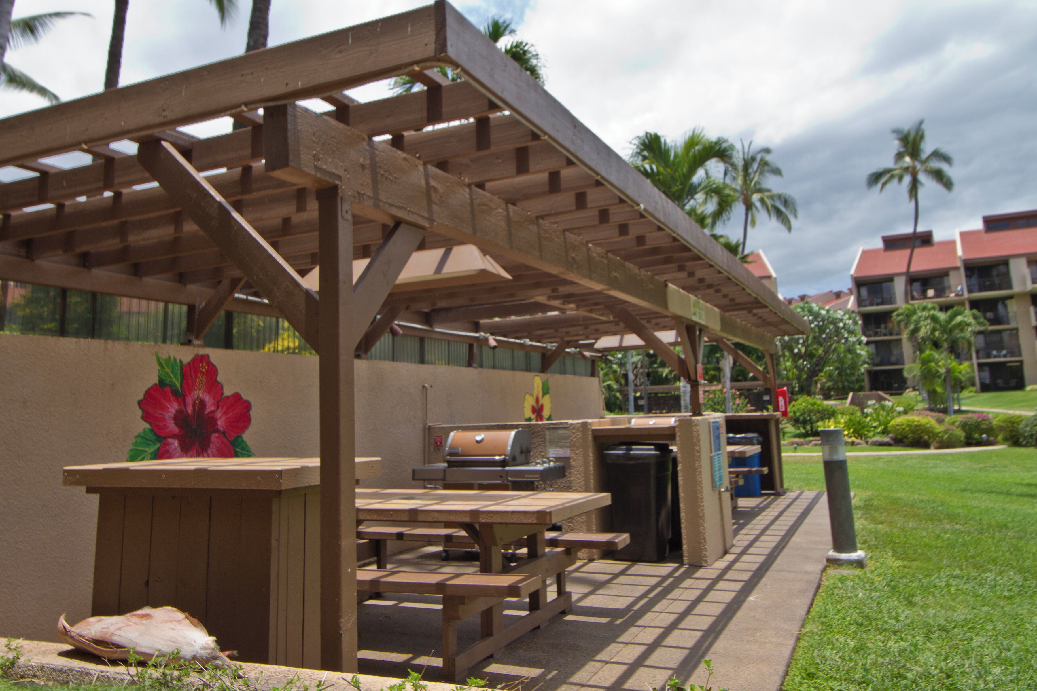 The outdoor barbecue pavilion features grills and picnic tables, surrounded by tropical gardens and palm trees under a wooden pergola—ideal for gathering.