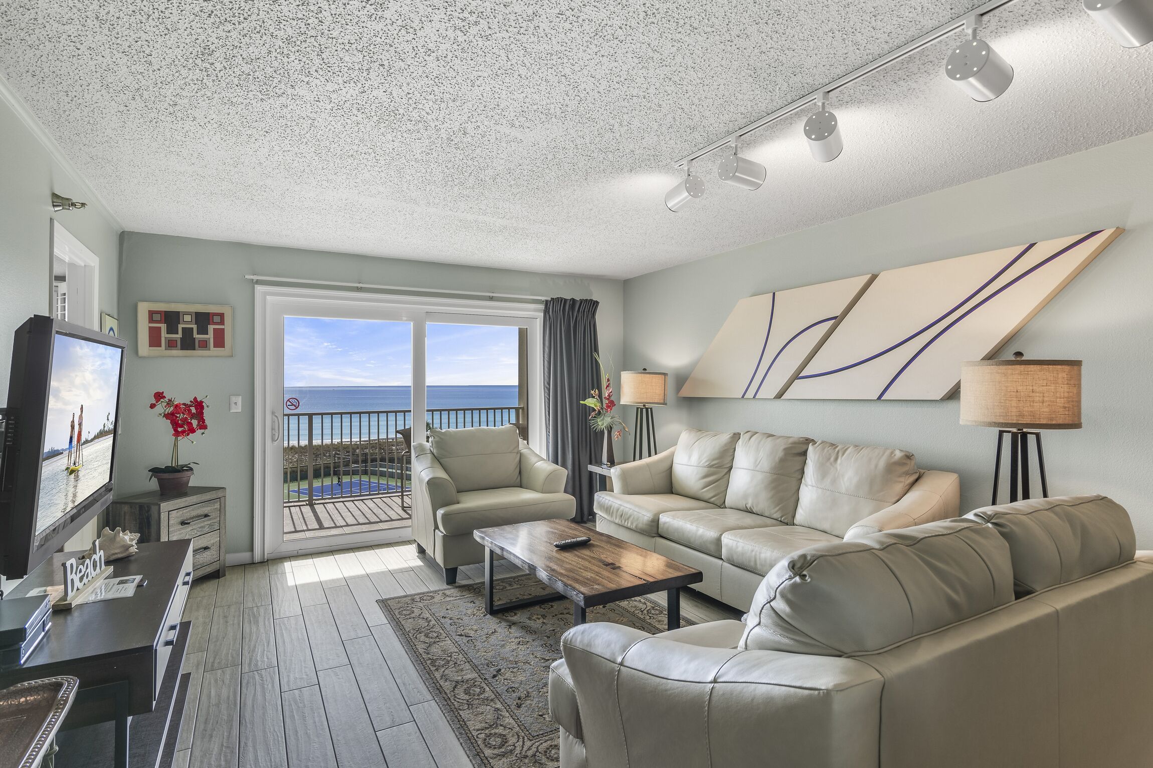 A living room with a TV, a coffee table, two sofas, and a balcony with a view of the ocean.