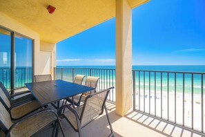 Un balcon avec une table et des chaises, donnant sur une plage où l’eau est d’un bleu limpide et le rivage est sablonneux.