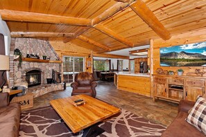 A cozy cabin interior with a stone fireplace, wooden furniture, and a large window overlooking a mountainous landscape.
