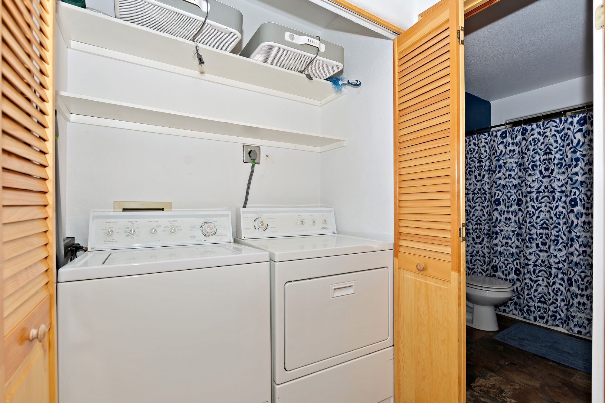 A laundry room with two stacked washers and dryers, a shelf above with a hairdryer and brush, and a toilet visible through an open door.