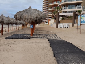 Une zone de plage avec des parasols en chaume, un panneau indiquant qu’elle est accessible aux personnes en fauteuil roulant, et un bâtiment doté de plusieurs balcons.