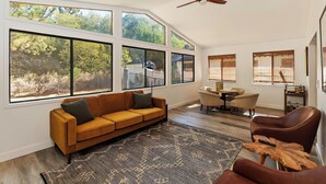 A spacious living room with a large window, a ceiling fan, a sofa, a coffee table, and a dining area. - Rise and Shine by Destination Big Bear : Pool Table! Shuffleboard! Walk to the Lake! Near Snow Summit! Internet! (Big bear lake)