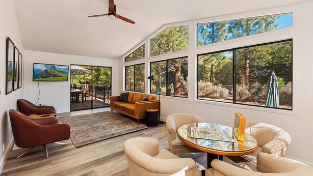 A spacious living room with large windows, a ceiling fan, and a round coffee table with books. - Rise and Shine by Destination Big Bear : Pool Table! Shuffleboard! Walk to the Lake! Near Snow Summit! Internet! (Big bear lake)