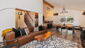 A spacious living room with a brown leather sofa, a wooden coffee table, and a dining area with a wooden table and chairs. A kitchen with a built-in stove and bar stools is visible in the background. - Rise and Shine by Destination Big Bear : Pool Table! Shuffleboard! Walk to the Lake! Near Snow Summit! Internet! (Big bear lake)