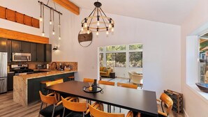 A modern kitchen with a central island, a dining area with a large table and chairs, and a view of the outdoors through large windows. - Rise and Shine by Destination Big Bear : Pool Table! Shuffleboard! Walk to the Lake! Near Snow Summit! Internet! (Big bear lake)