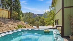 A hot tub in a backyard with a mountain view, surrounded by trees and a wooden deck. - Movie Theater! Pool Table! HOT TUB! Mtn Views! Multiple Decks! (Big Bear Lake)