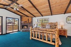 A bedroom with a large bed, a fireplace, a ceiling fan, and a view of the outdoors.