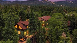 A two-story cabin with a deck, surrounded by dense forest and mountains in the background. - Movie Theater! Pool Table! HOT TUB! Mtn Views! Multiple Decks! (Big Bear Lake)