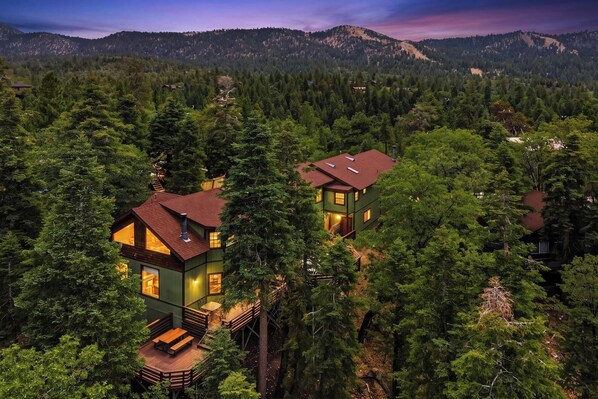 A two-story cabin with a deck, surrounded by dense forest and mountains in the background.