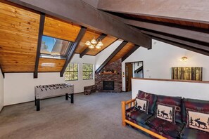 Un espacio tipo loft con techo de madera, chimenea, sofá con cojines de motivos animales y una mesa de futbolín.