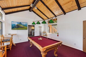 A pool table in a room with a high ceiling and wooden beams, a painting of a landscape, and a dining area with chairs and a table.