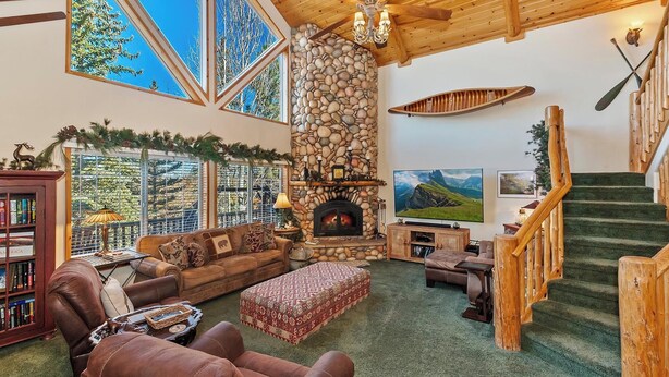 Una sala de estar acogedora con chimenea de piedra, ventanas amplias, una escalera de madera y una estantería.