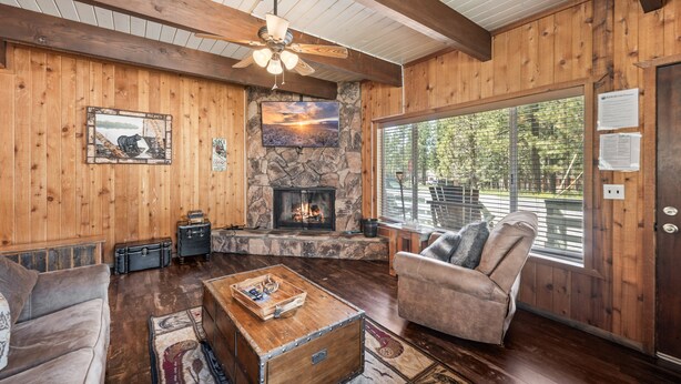 A wooden interior with a fireplace, a ceiling fan, and a view through a large window.
