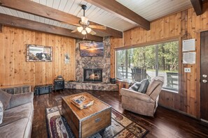 A wooden interior with a fireplace, a ceiling fan, and a view through a large window.
