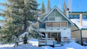 A two-story lodge with a balcony, surrounded by snow-covered trees and a wooden deck.