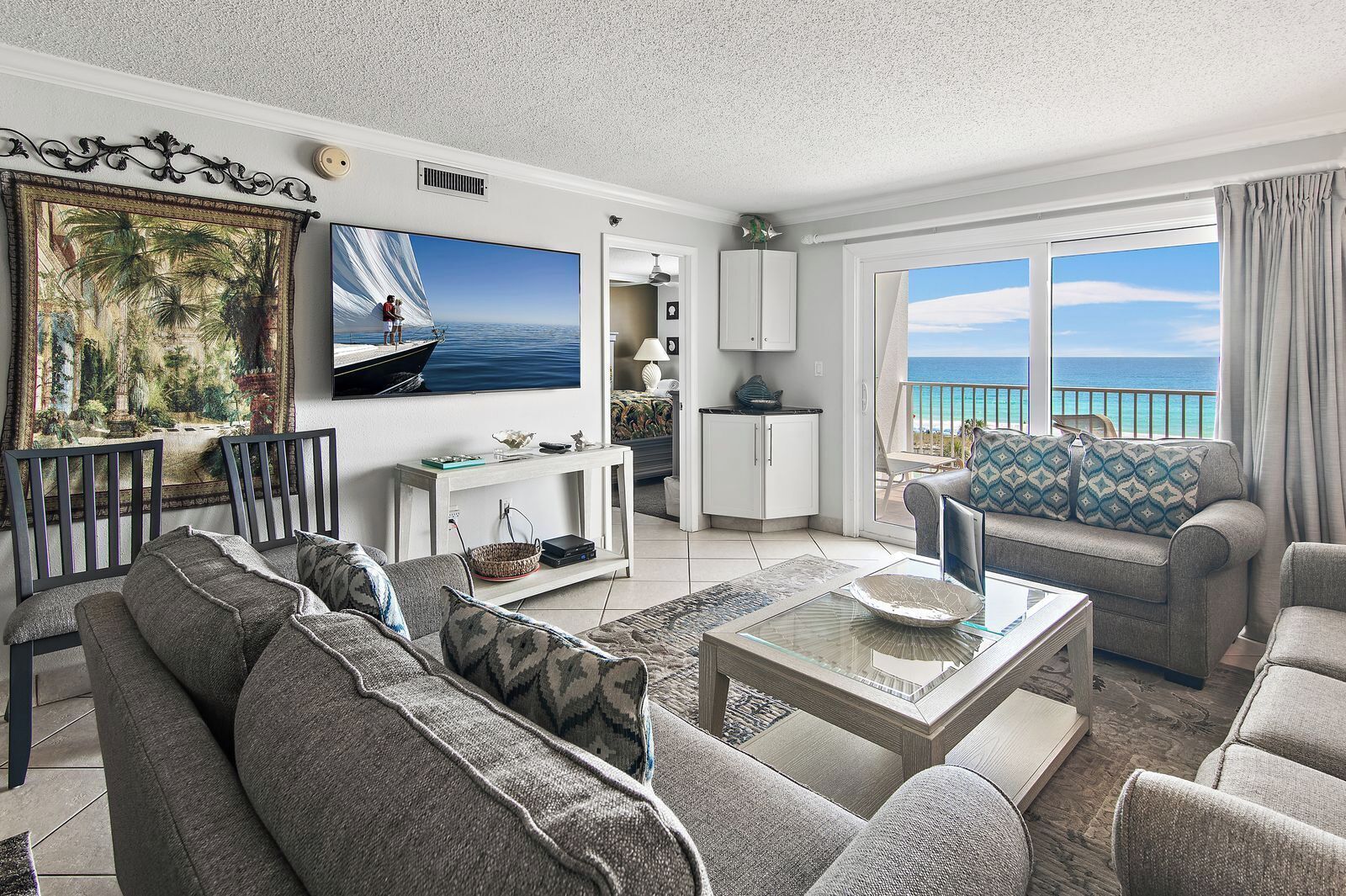 A spacious living room with a large TV, comfortable seating, a dining area, and a balcony with a view of the ocean.