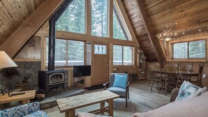 En mysig stuga med en stor öppen spis, trämöbler och stora fönster som ger utsikt över skogen.