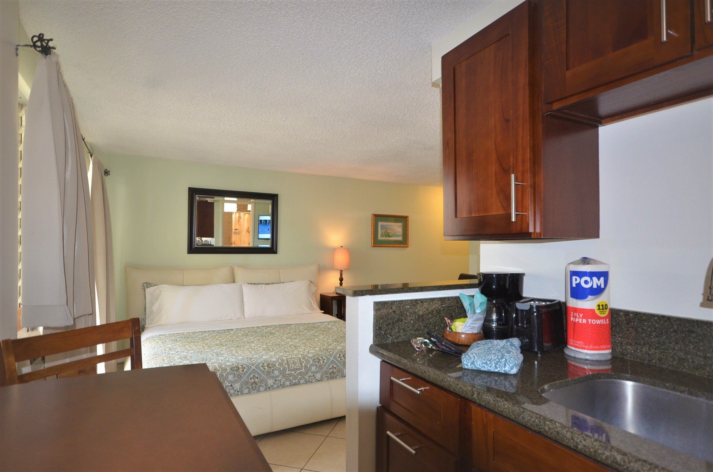 A compact hotel room with a bed, a small kitchenette, a dining table, and a bathroom with a shower curtain.