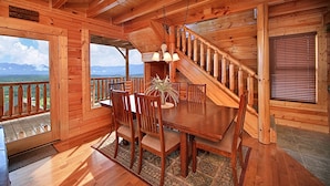 A wooden cabin interior with a dining area, a staircase, and a view of mountains.