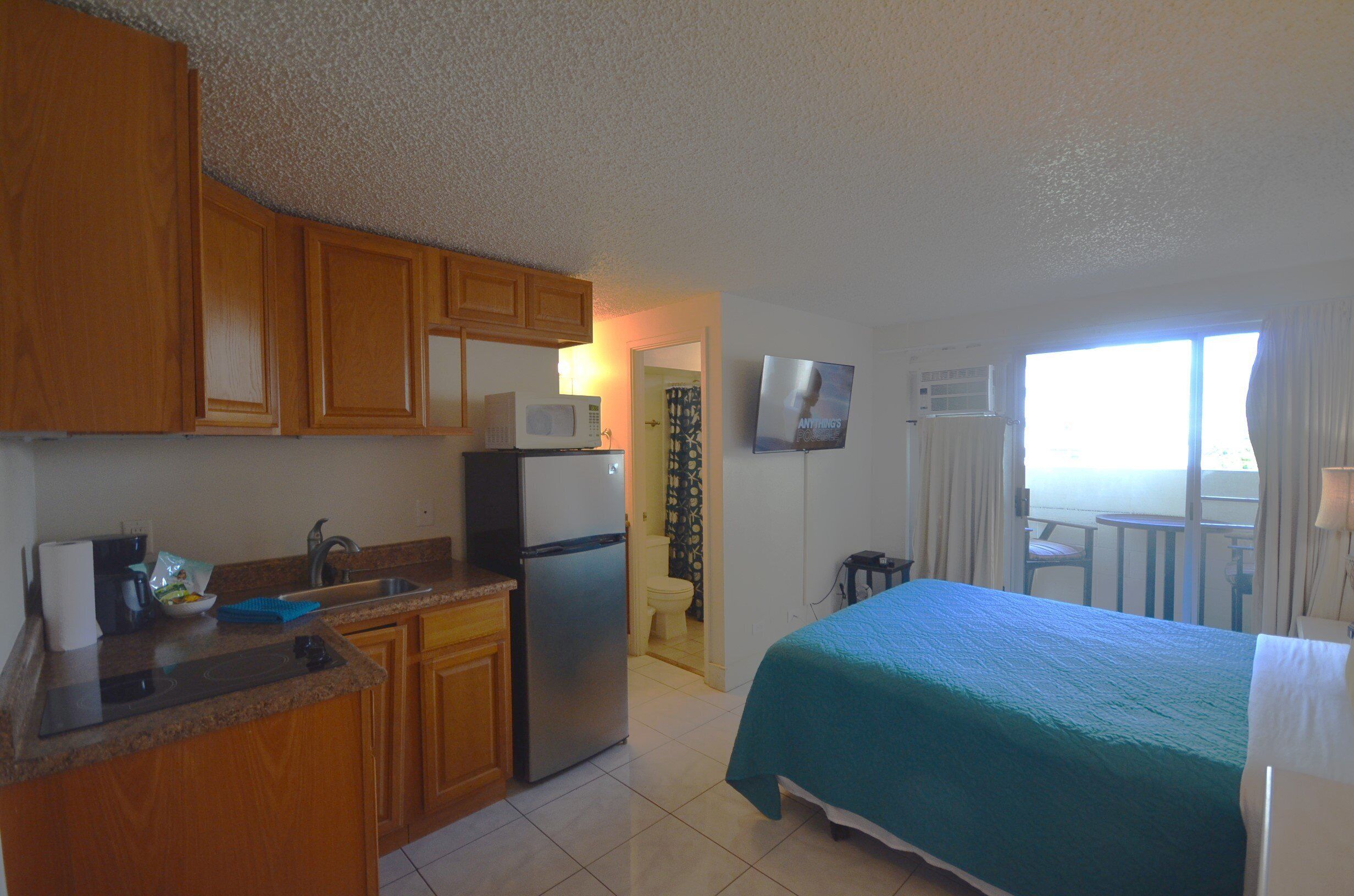 A compact hotel room with a kitchenette, a bed with a teal bedspread, a television mounted on the wall, and a balcony with a view.