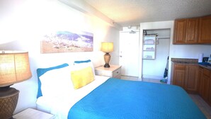 A bedroom with a bed, bedside lamps, a nightstand, a painting, and a view of the kitchen.