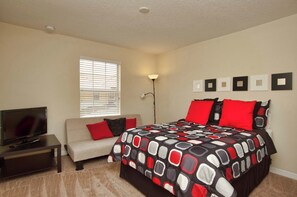 A bedroom with a bed, a television on a stand, a sofa, and a window with blinds.