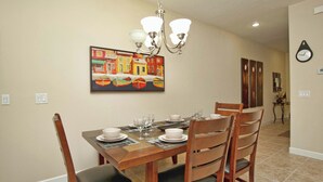 Uma sala de jantar com mesa de madeira posta para quatro pessoas, cadeiras, um lustre e um quadro de barcos na parede.
