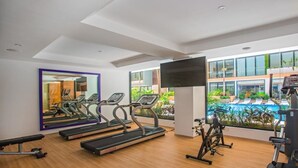 Ett gym med löpband, en motionscykel och en träningsbänk. Det finns en stor spegel på väggen och en tv som är monterad på väggen. Gymmet har stora fönster med utsikt över en pool och grönområden utanför.