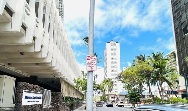 A street view with a 'NO PARKING 3 MINUTE PASSENGER LOADING ZONE' sign, a building with a sign for 'Marine Surf Waikiki', and a tall building in the background.
