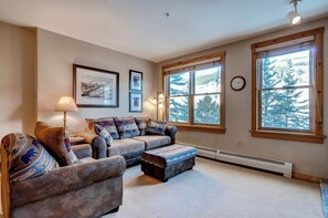 A cozy living room with a sofa, ottoman, and a large window with a view of trees. - Walk To The Slopes! Full kitchen Sleeps 4 (Keystone)