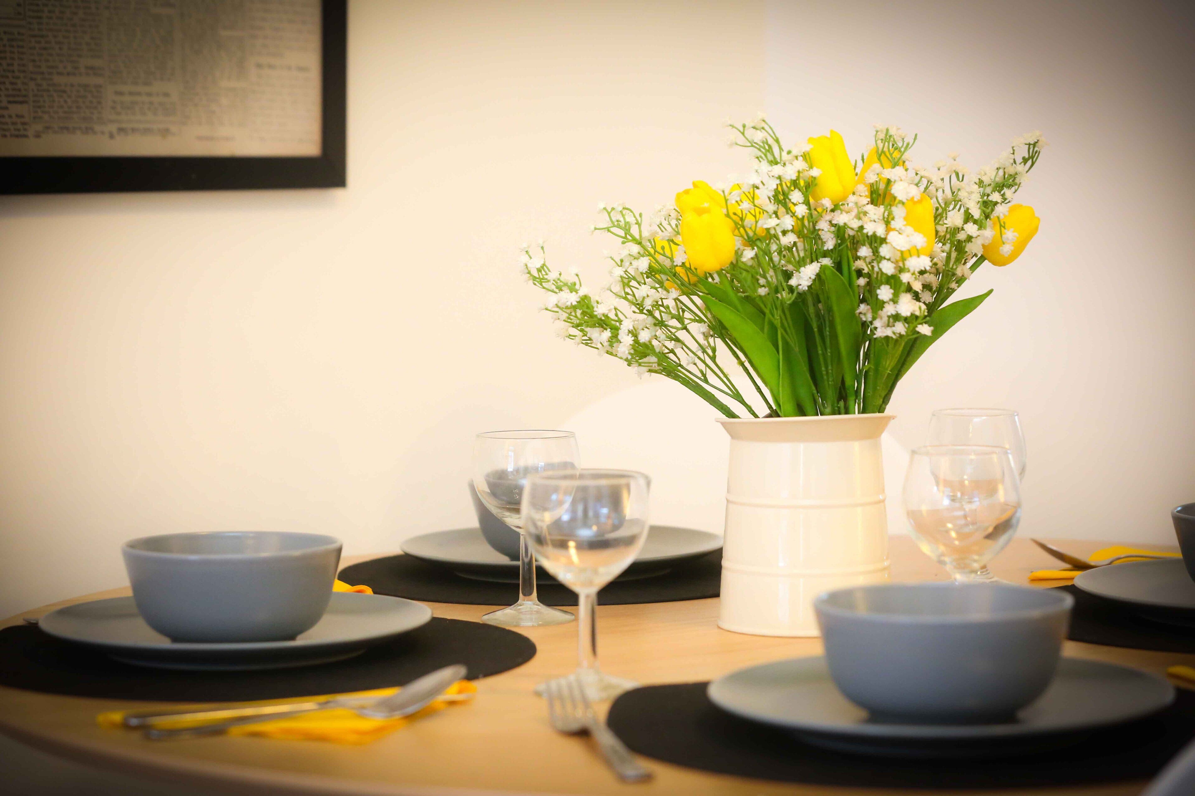 Une table à manger dressée avec des assiettes, des bols, des couverts et un vase rempli de fleurs jaunes.